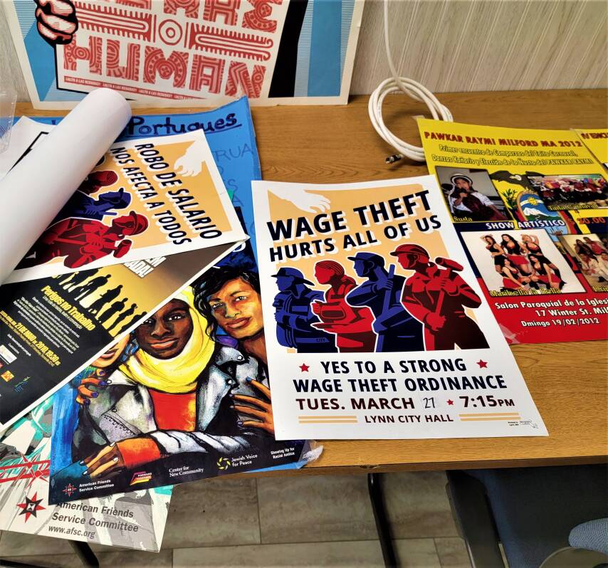 Several posters overlap on the top of a table. They all show illustrations of people, and the words of the frontmost poster read "Wage theft hurts all of us."