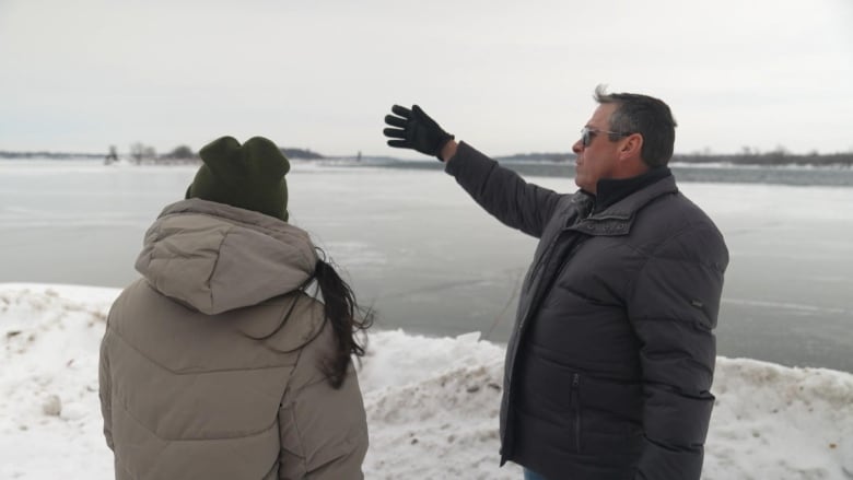 A man gestures toward a waterway in winter with a reporter next to him.