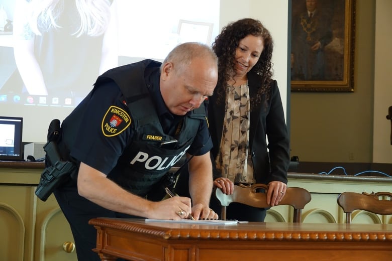 A middle-aged man wearing a police uniform bends over a low table to sign a piece of paper. Lana Saunders, who has lots of dark, curly hair, stands next to him, smiling.