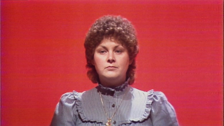 Film still from a 1980s TV program To Tell The Truth. Shows a medium closeup of Michelle Smith, a white woman with a short brown perm. She wears a high-collar microfloral dress and looks at the camera with blank expression. The background is red.